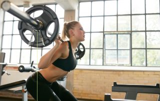 Young woman athlete exercising using barbell with heavy weights in a gym. Measuring her athletic performance with Tendo Unit by Tendo Sport to make her weight training efficient