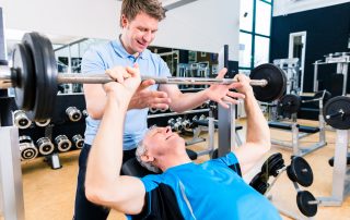 Trainer assisting senior man lifting barbell in gym while measuring his athletic performance via Tendo Unit by Tendo Sport to safely gain strength and fitness