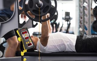 Man athlete doing barbell bench press in gym with personal trainer while measuring his velocity for velocity based training which is shown on a display of Tendo microcomputer unit by Tendo Sport