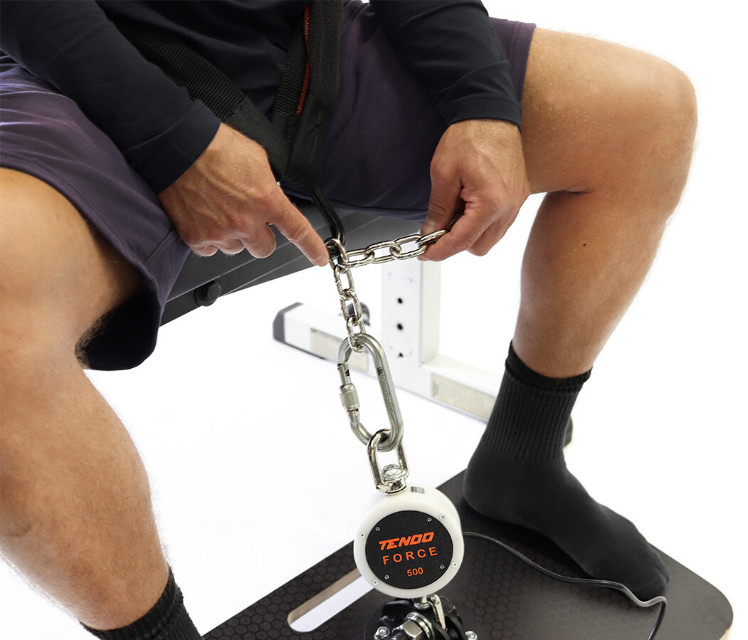 Tendo Force – setup Athlete sitting on a bench is clipping his waist belt into a chain that is attached onto the Tendo Force sensor while both his feet are placed on the inclined platform ready to perform isometric belt squat